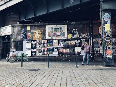 An urban street scene with a graffiti-covered wall displaying a variety of posters and street art beneath an overpass. A man stands beside the wall holding a drink, while a few bicycles are parked nearby. The atmosphere is gritty and artistic, reflecting a vibrant urban culture.