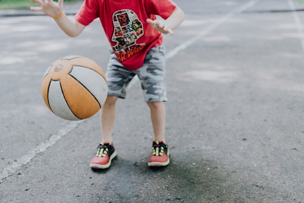 A child in a red shirt and camouflage shorts is playing with a basketball on a pavement. The basketball is orange and white, captured mid-bounce, while the child's hands are positioned to catch or dribble it. The child is wearing red sneakers with neon green details.