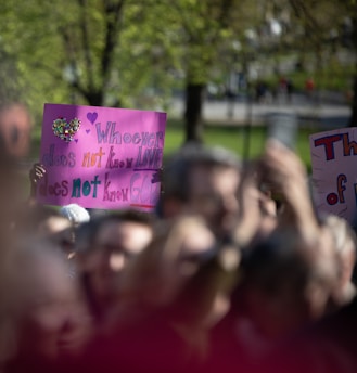 A group of people holding colorful signs during a gathering or protest in a park setting. The forefront sign reads 'Whoever does not know love does not know God' written with decorative letters including a heart embellishment. The crowd appears blurred, indicating movement or focus on the signs.