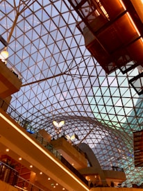 A geometric glass ceiling with a grid pattern creates an open and spacious interior. Multiple floors are visible with warm lighting and modern architecture, including railings and plant installations. The ceiling's structure allows natural light, enhancing the modern aesthetic.
