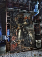 A large, intricate painting is displayed within a construction area, surrounded by scaffolding. The artwork features a religious scene with multiple figures, including a central bishop in ornate ecclesiastical attire with a mitre, and several other individuals in various traditional garments. At the top, there is a depiction of the Virgin Mary holding the Christ Child, surrounded by cherubs. The surface of the painting appears aged and weathered.