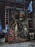 A large, intricate painting is displayed within a construction area, surrounded by scaffolding. The artwork features a religious scene with multiple figures, including a central bishop in ornate ecclesiastical attire with a mitre, and several other individuals in various traditional garments. At the top, there is a depiction of the Virgin Mary holding the Christ Child, surrounded by cherubs. The surface of the painting appears aged and weathered.
