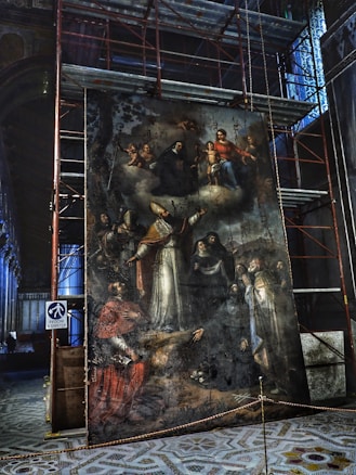 A large, intricate painting is displayed within a construction area, surrounded by scaffolding. The artwork features a religious scene with multiple figures, including a central bishop in ornate ecclesiastical attire with a mitre, and several other individuals in various traditional garments. At the top, there is a depiction of the Virgin Mary holding the Christ Child, surrounded by cherubs. The surface of the painting appears aged and weathered.