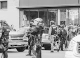 Police officers in tactical gear, including helmets and body armor, stand on a city street. Several officers carry batons and other equipment. Behind them, there are civilians observing and capturing the scene with smartphones. Vehicles are parked along the street, and a building with large windows is visible in the background.