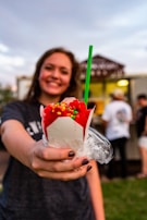 A person holds out a bright red shaved ice in a white cup topped with colorful candy sprinkles and a green straw. The background is slightly blurred, with other people and a food truck visible.