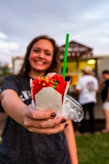 A person holds out a bright red shaved ice in a white cup topped with colorful candy sprinkles and a green straw. The background is slightly blurred, with other people and a food truck visible.
