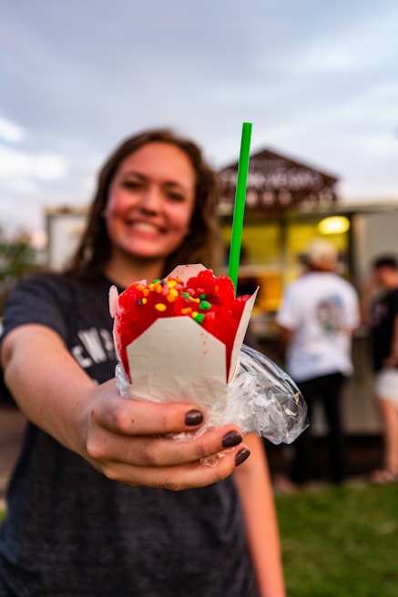 A person holds out a bright red shaved ice in a white cup topped with colorful candy sprinkles and a green straw. The background is slightly blurred, with other people and a food truck visible.