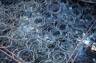 A collection of metal bed springs in a worn and rusty condition, laying on a dark, ashy surface with signs of charring and burn marks on the surrounding area.