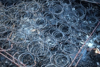 A collection of metal bed springs in a worn and rusty condition, laying on a dark, ashy surface with signs of charring and burn marks on the surrounding area.