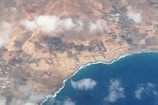 An aerial view of a coastal region featuring a deep blue ocean along the shore. The land is predominantly arid with reddish-brown tones and geometric patterns indicating agricultural fields or plots. There are clouds casting shadows over the landscape, and small clusters of buildings scattered around the area.
