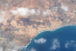 An aerial view of a coastal region featuring a deep blue ocean along the shore. The land is predominantly arid with reddish-brown tones and geometric patterns indicating agricultural fields or plots. There are clouds casting shadows over the landscape, and small clusters of buildings scattered around the area.