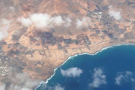 An aerial view of a coastal region featuring a deep blue ocean along the shore. The land is predominantly arid with reddish-brown tones and geometric patterns indicating agricultural fields or plots. There are clouds casting shadows over the landscape, and small clusters of buildings scattered around the area.
