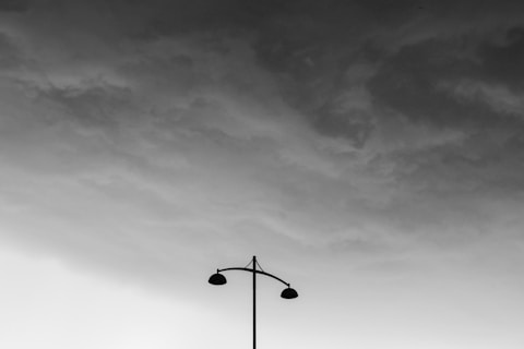 Dark, overcast sky with heavy, textured clouds looming above a solitary streetlamp. The scene is in grayscale, emphasizing the dramatic contrast between the stormy sky and the silhouetted lamp.