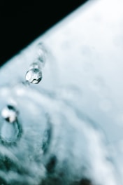 A close-up of a water droplet suspended in mid-air, with a blurred background that includes more water droplets. The image captures the clarity and translucence of the droplet.