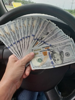 A hand holding cash with a scrap car in the background.