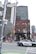 A street view featuring various buildings, including a red-brick building with multiple arched windows and taller modern buildings in the background. A police car is driving through the intersection with pedestrians in the vicinity. The street sign reads 'Queen St. W.' in both English and Chinese characters.