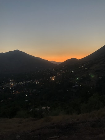 The image captures a view of a small town or village nestled in a valley as the sun sets behind surrounding mountains. The sky transitions from a soft orange near the horizon to a deep blue above, creating a serene backdrop. Lights from the houses dot the landscape, adding a warm and cozy atmosphere to the scene.