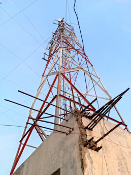 A tall metal telecommunications tower with a lattice structure rises against a clear sky. It is supported by several steel beams and cables and is mounted on a concrete base. Visible corrosion and wear mark the structure, with some broken beams protruding from the concrete.
