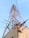 A tall metal telecommunications tower with a lattice structure rises against a clear sky. It is supported by several steel beams and cables and is mounted on a concrete base. Visible corrosion and wear mark the structure, with some broken beams protruding from the concrete.