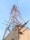 A tall metal telecommunications tower with a lattice structure rises against a clear sky. It is supported by several steel beams and cables and is mounted on a concrete base. Visible corrosion and wear mark the structure, with some broken beams protruding from the concrete.