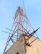 A tall metal telecommunications tower with a lattice structure rises against a clear sky. It is supported by several steel beams and cables and is mounted on a concrete base. Visible corrosion and wear mark the structure, with some broken beams protruding from the concrete.