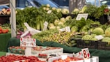 A vibrant market stall displays a variety of fresh fruits and vegetables, including strawberries, watermelons, zucchinis, carrots, and leafy greens. Price signs are visible, indicating deals and prices for different produce. The produce is neatly arranged in baskets and crates on a green tablecloth.