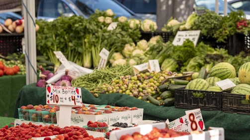 A vibrant market stall displays a variety of fresh fruits and vegetables, including strawberries, watermelons, zucchinis, carrots, and leafy greens. Price signs are visible, indicating deals and prices for different produce. The produce is neatly arranged in baskets and crates on a green tablecloth.