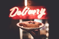 A hand holds a cake on a decorative plate in front of a brightly lit 'Delivery' sign. The cake appears to be a cheesecake, topped with some red fruit or sauce garnish.