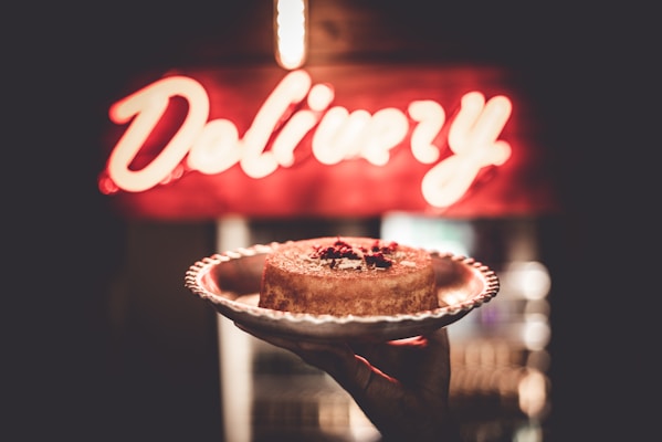 A hand holds a cake on a decorative plate in front of a brightly lit 'Delivery' sign. The cake appears to be a cheesecake, topped with some red fruit or sauce garnish.
