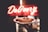 A hand holds a cake on a decorative plate in front of a brightly lit 'Delivery' sign. The cake appears to be a cheesecake, topped with some red fruit or sauce garnish.