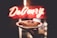 A hand holds a cake on a decorative plate in front of a brightly lit 'Delivery' sign. The cake appears to be a cheesecake, topped with some red fruit or sauce garnish.