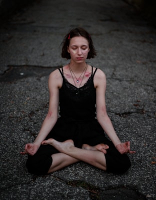 A person sitting cross-legged in a meditative pose on a rough, textured surface. They are wearing a black sleeveless top and black pants. Their eyes are closed, and they have a calm, focused expression.