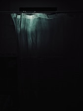 A shower curtain hangs in a dimly lit bathroom. The upper part of the curtain is highlighted by a faint light, creating a soft contrast with the surrounding darkness. The curtain appears slightly wrinkled with a textured pattern on the lower section.