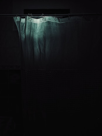 A shower curtain hangs in a dimly lit bathroom. The upper part of the curtain is highlighted by a faint light, creating a soft contrast with the surrounding darkness. The curtain appears slightly wrinkled with a textured pattern on the lower section.