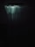 A shower curtain hangs in a dimly lit bathroom. The upper part of the curtain is highlighted by a faint light, creating a soft contrast with the surrounding darkness. The curtain appears slightly wrinkled with a textured pattern on the lower section.