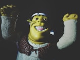 A cartoon character figure with green skin and a wide, toothy grin is wearing a white headband and a textured brown and beige outfit. The character is raising its fists in a triumphant pose, creating a playful and energetic atmosphere. The lighting emphasizes the character's features against a dark background.
