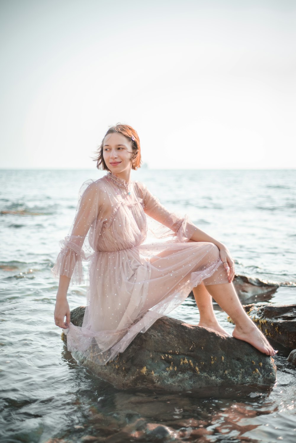 woman wearing beige long-sleeved dress sitting on rock while glancing her right side near sea during daytime