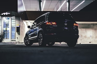 A dark, sleek SUV is parked in an illuminated urban setting, possibly an underground garage. The car's taillights are on, casting a soft red glow. Architectural details include a modern lighted ceiling with lines and concrete walls. An ATM or similar structure is visible to the left.