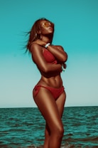 A woman stands confidently at the beach, wearing a red bikini and sunglasses. The ocean and sky create a serene backdrop, and her arms are crossed around her chest, conveying a feeling of relaxation and empowerment.