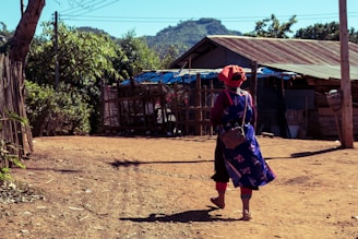 A person wearing traditional attire with a vibrant and patterned dress is walking along a dirt path in a rural setting. The scene includes wooden structures with tin roofs, trees with green foliage, and distant hills under a clear blue sky.
