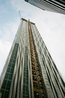 A tall building under construction extends upwards into the sky, creating a sense of height and scale. It is framed by two other skyscrapers, one on each side. The building features a crane at the top and has a facade of glass and metal.