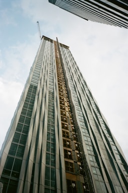 A tall building under construction extends upwards into the sky, creating a sense of height and scale. It is framed by two other skyscrapers, one on each side. The building features a crane at the top and has a facade of glass and metal.