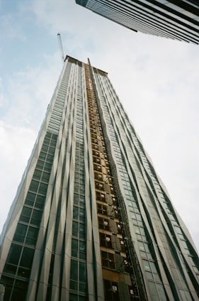 A tall building under construction extends upwards into the sky, creating a sense of height and scale. It is framed by two other skyscrapers, one on each side. The building features a crane at the top and has a facade of glass and metal.