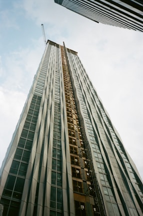 A tall building under construction extends upwards into the sky, creating a sense of height and scale. It is framed by two other skyscrapers, one on each side. The building features a crane at the top and has a facade of glass and metal.