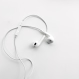 A pair of white wired earphones with a built-in control module rests on a flat, clean white surface. The earphones are neatly arranged, creating a minimalist and modern aesthetic.
