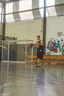 A person wearing a basketball jersey is playing with a ball inside an indoor sports court. The background features a brick wall with colorful graffiti art and a wire fence partially surrounding the area.