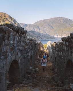 A pathway framed by ancient stone walls leads towards a picturesque view of a bay flanked by rugged hills. Two people walk along the path, one carrying a backpack. The scene is bathed in natural light, highlighting the textures of the stone and the serene landscape in the background.