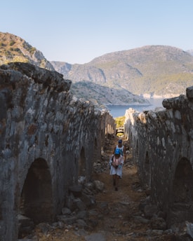 A pathway framed by ancient stone walls leads towards a picturesque view of a bay flanked by rugged hills. Two people walk along the path, one carrying a backpack. The scene is bathed in natural light, highlighting the textures of the stone and the serene landscape in the background.