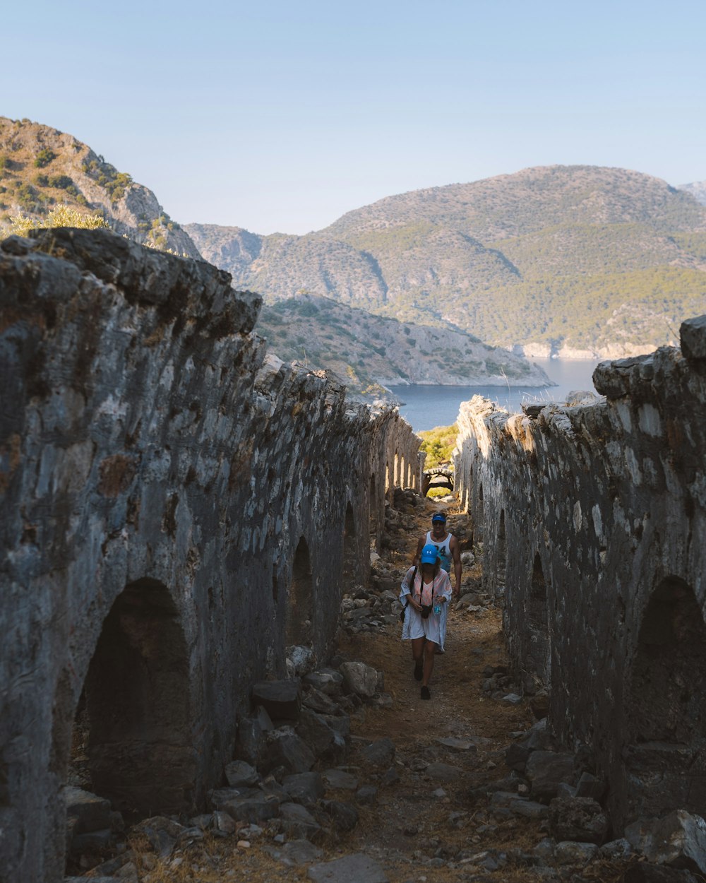 two people walking near bridge viewing mountain and body of water during daytime