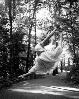 A striking black-and-white photo of a dancer caught mid-leap, set against a softly blurred backdrop of towering Pacific Northwest trees.